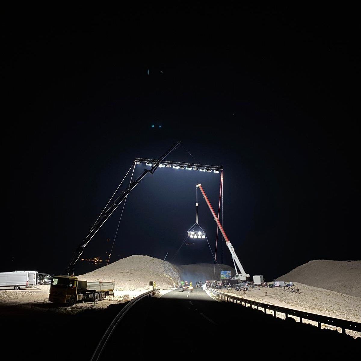 Two cranes flying a lighting truss over a sand quarry at night