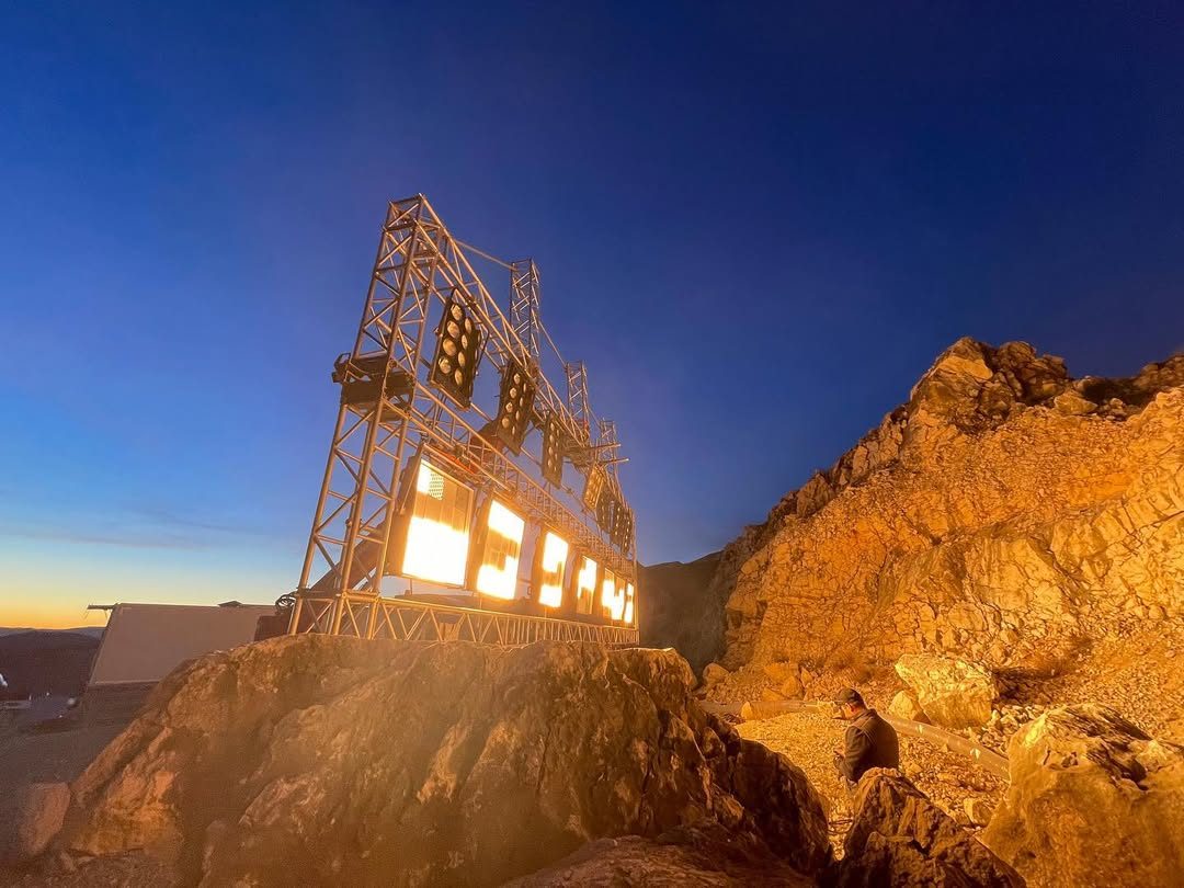 Lit window-frame rig on scaffolding on a rocky ridge at dusk