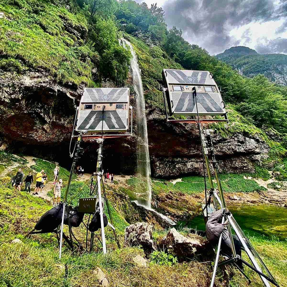 Two large lighting panels on stands beside a waterfall in a green gorge